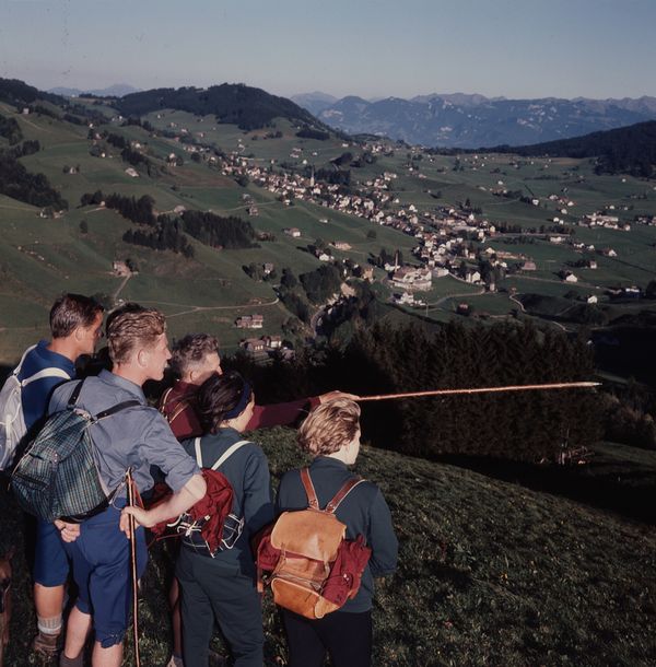 Bild einer Wandergruppe "in blauen Kleidern" die von einem Hügel in die Ferne schauen. Eine Person zeigt etwas mit einem Wanderstock. 